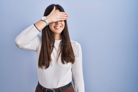 Photo for Young brunette woman standing over blue background smiling and laughing with hand on face covering eyes for surprise. blind concept. - Royalty Free Image