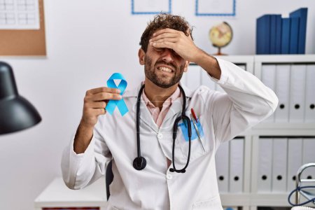 Téléchargez les photos : Young hispanic doctor man holding blue ribbon stressed and frustrated with hand on head, surprised and angry face - en image libre de droit