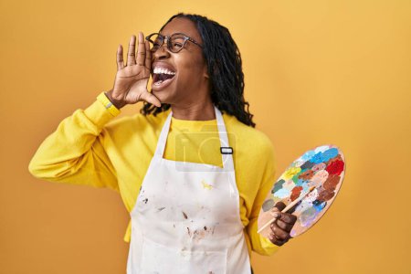 Téléchargez les photos : African woman holding painter palette shouting and screaming loud to side with hand on mouth. communication concept. - en image libre de droit