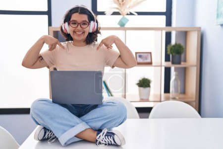 Photo for Young hispanic woman using laptop sitting on the table wearing headphones looking confident with smile on face, pointing oneself with fingers proud and happy. - Royalty Free Image