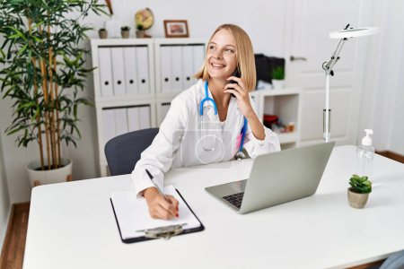 Foto de Mujer rubia joven vistiendo uniforme médico hablando en el teléfono inteligente en la clínica - Imagen libre de derechos