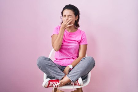 Foto de Hispanic young woman sitting on chair over pink background smelling something stinky and disgusting, intolerable smell, holding breath with fingers on nose. bad smell - Imagen libre de derechos