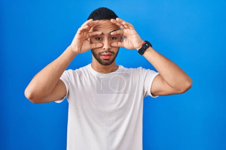 Téléchargez les photos : Young hispanic man standing over blue background trying to open eyes with fingers, sleepy and tired for morning fatigue - en image libre de droit