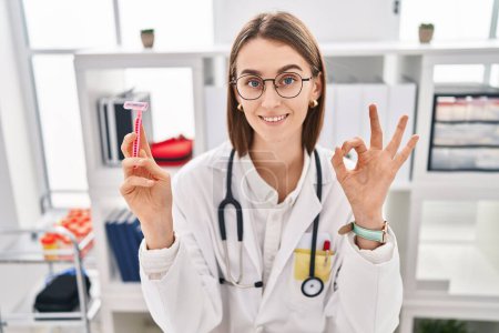 Téléchargez les photos : Young caucasian doctor woman holding razor doing ok sign with fingers, smiling friendly gesturing excellent symbol - en image libre de droit