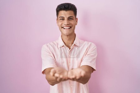 Foto de Young hispanic man standing over pink background smiling with hands palms together receiving or giving gesture. hold and protection - Imagen libre de derechos