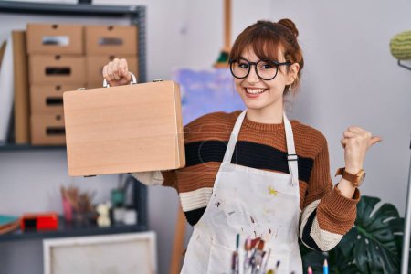 Téléchargez les photos : Jeune belle artiste femme tenant boîtier en bois pointant pouce vers le côté souriant heureux avec bouche ouverte - en image libre de droit