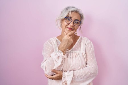 Photo for Middle age woman with grey hair standing over pink background looking confident at the camera with smile with crossed arms and hand raised on chin. thinking positive. - Royalty Free Image