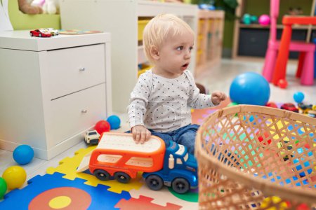 Téléchargez les photos : Adorable tout-petit blond jouant avec des voitures jouet assis sur le sol à la maternelle - en image libre de droit