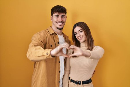 Téléchargez les photos : Young hispanic couple standing over yellow background smiling in love doing heart symbol shape with hands. romantic concept. - en image libre de droit
