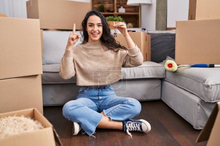Téléchargez les photos : Young brunette woman sitting on the floor at new home holding keys surprised with an idea or question pointing finger with happy face, number one - en image libre de droit