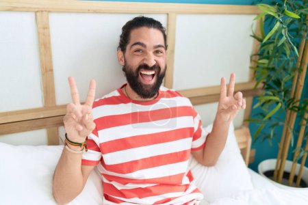 Foto de Joven hombre hispano haciendo gesto de victoria con los dedos sentados en la cama en el dormitorio - Imagen libre de derechos