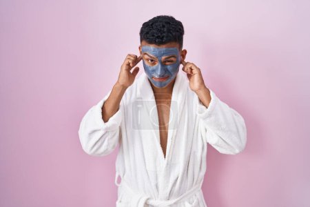 Téléchargez les photos : Young hispanic man wearing beauty face mask and bath robe covering ears with fingers with annoyed expression for the noise of loud music. deaf concept. - en image libre de droit