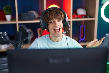 Téléchargez les photos : Jeune homme hispanique jouant à des jeux vidéo célébrant la réalisation avec un sourire heureux et l'expression gagnante avec la main levée - en image libre de droit