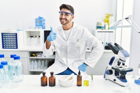 Foto de Young hispanic man working at scientist laboratory smiling happy and positive, thumb up doing excellent and approval sign - Imagen libre de derechos