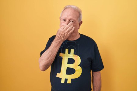 Téléchargez les photos : Senior man with grey hair wearing bitcoin t shirt smelling something stinky and disgusting, intolerable smell, holding breath with fingers on nose. bad smell - en image libre de droit