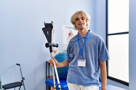 Foto de Joven hombre rubio pisioterapeuta sonriendo confiado sosteniendo muletas en la clínica de rehabilitación - Imagen libre de derechos