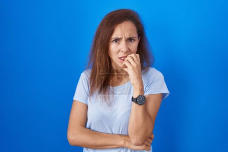 Foto de Brunette woman standing over blue background looking stressed and nervous with hands on mouth biting nails. anxiety problem. - Imagen libre de derechos