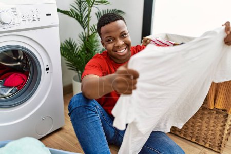Foto de Hombre africano joven mirando camiseta fresca y limpia en la sala de lavandería - Imagen libre de derechos