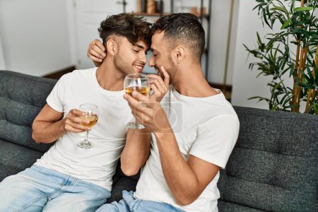 Foto de Dos hombres hispanos brindando en pareja con una copa de vino sentados en un sofá en casa - Imagen libre de derechos