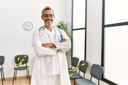 Foto de Médico hombre de pelo gris de mediana edad sonriendo confiado de pie con los brazos cruzados gesto en la sala de espera clínica - Imagen libre de derechos