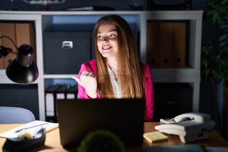 Photo for Young caucasian woman working at the office at night smiling with happy face looking and pointing to the side with thumb up. - Royalty Free Image