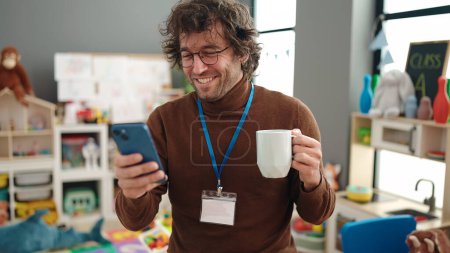 Téléchargez les photos : Young hispanic man preschool teacher using smartphone drinking coffee at kindergarten - en image libre de droit