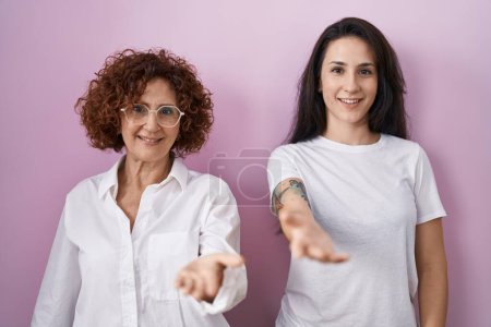Photo for Hispanic mother and daughter wearing casual white t shirt over pink background smiling cheerful offering palm hand giving assistance and acceptance. - Royalty Free Image