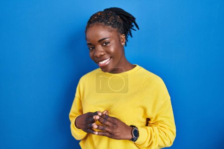 Téléchargez les photos : Beautiful black woman standing over blue background hands together and fingers crossed smiling relaxed and cheerful. success and optimistic - en image libre de droit