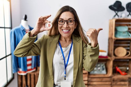 Téléchargez les photos : Jeune femme hispanique tenant Bitcoin travaillant au magasin de vêtements pointant pouce vers le côté souriant heureux avec la bouche ouverte - en image libre de droit