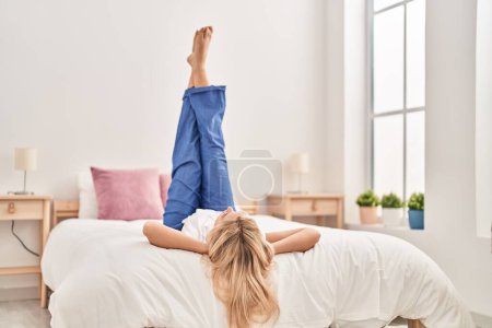 Téléchargez les photos : Jeune femme blonde allongée sur le lit avec les jambes levées dans la chambre - en image libre de droit