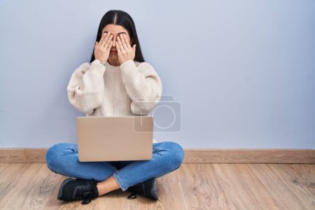 Foto de Mujer joven usando portátil sentado en el suelo en casa frotando los ojos para la fatiga y el dolor de cabeza, la expresión somnolienta y cansada. problema de visión - Imagen libre de derechos