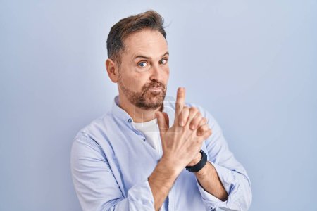 Téléchargez les photos : Middle age caucasian man standing over blue background holding symbolic gun with hand gesture, playing killing shooting weapons, angry face - en image libre de droit