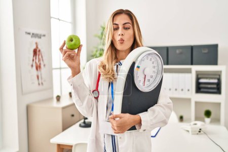 Foto de Young blonde doctor woman holding weighing machine and green apple making fish face with mouth and squinting eyes, crazy and comical. - Imagen libre de derechos
