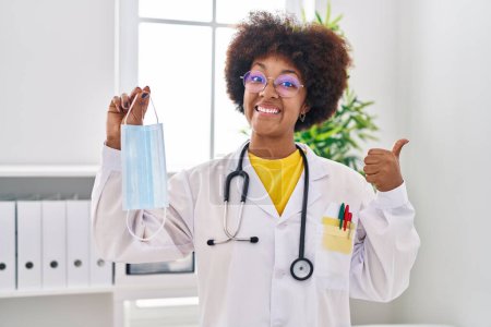 Foto de Young african american woman wearing doctor uniform holding safety mask pointing thumb up to the side smiling happy with open mouth - Imagen libre de derechos