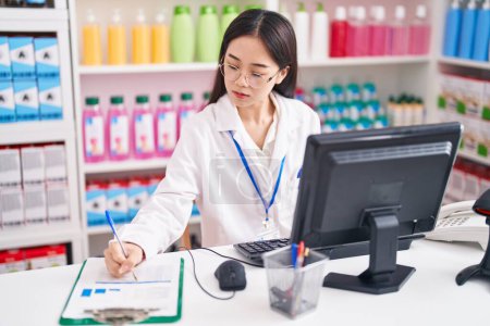 Photo for Young chinese woman pharmacist writing on document using computer at pharmacy - Royalty Free Image