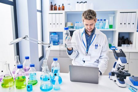 Téléchargez les photos : Jeune homme scientifique à l'aide d'un ordinateur portable tenant bouteille au laboratoire - en image libre de droit