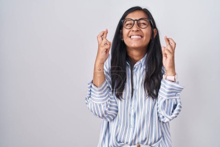 Téléchargez les photos : Young hispanic woman wearing glasses gesturing finger crossed smiling with hope and eyes closed. luck and superstitious concept. - en image libre de droit