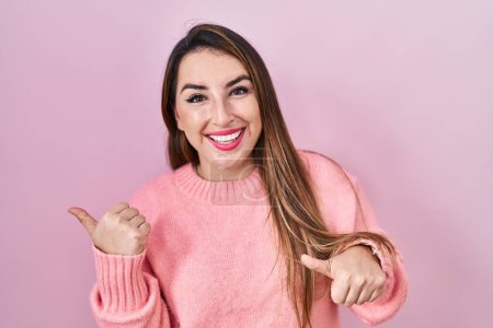 Foto de Young hispanic woman standing over pink background pointing to the back behind with hand and thumbs up, smiling confident - Imagen libre de derechos