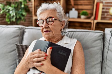 Foto de Mujer mayor de cabello gris abrazando la Biblia sentada en el sofá en casa - Imagen libre de derechos