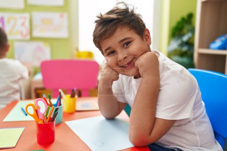 Téléchargez les photos : Adorable garçon hispanique étudiant souriant confiant assis sur la table à la maternelle - en image libre de droit