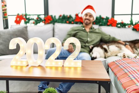 Téléchargez les photos : Jeune homme hispanique étreignant chien célébrer le nouvel an assis par décor de Noël à la maison - en image libre de droit