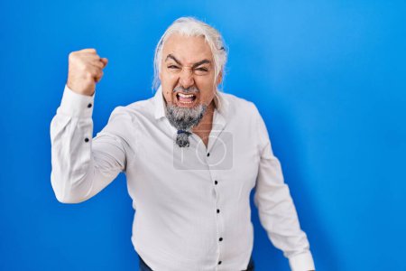 Téléchargez les photos : Homme du Moyen Âge avec les cheveux gris debout sur fond bleu en colère et fou levant le poing frustré et furieux tout en criant avec colère. rage et concept agressif. - en image libre de droit