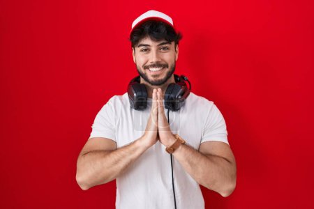 Téléchargez les photos : Hispanic man with beard wearing gamer hat and headphones praying with hands together asking for forgiveness smiling confident. - en image libre de droit