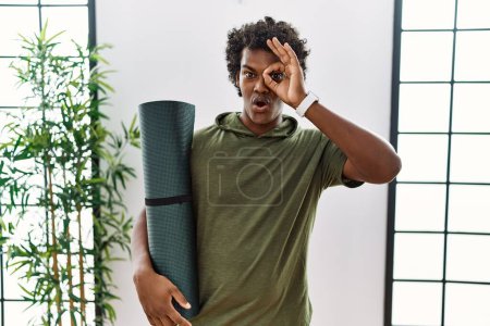 Téléchargez les photos : African man with curly hair holding yoga mat at studio doing ok gesture shocked with surprised face, eye looking through fingers. unbelieving expression. - en image libre de droit