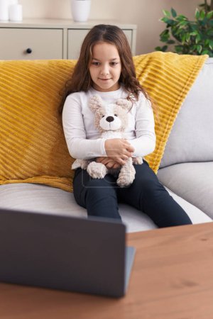 Foto de Adorable hispanic girl hugging teddy bear watching movie on laptop at home - Imagen libre de derechos