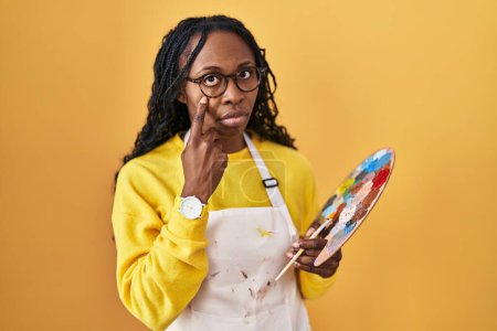 Téléchargez les photos : African woman holding painter palette pointing to the eye watching you gesture, suspicious expression - en image libre de droit