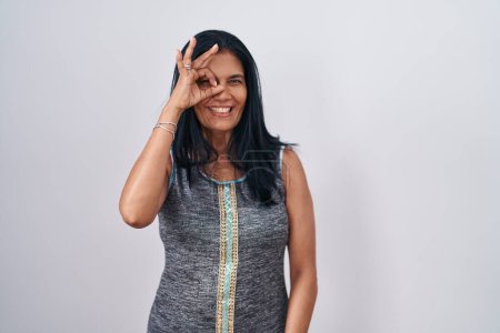 Foto de Mature hispanic woman standing over white background doing ok gesture with hand smiling, eye looking through fingers with happy face. - Imagen libre de derechos