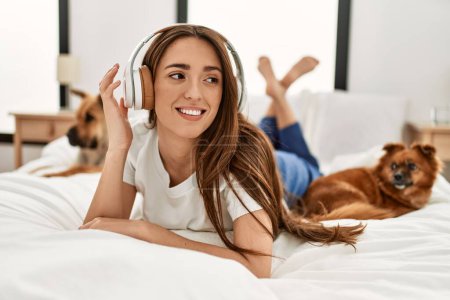 Téléchargez les photos : Jeune femme hispanique écoutant de la musique couchée sur le lit avec des chiens dans la chambre - en image libre de droit