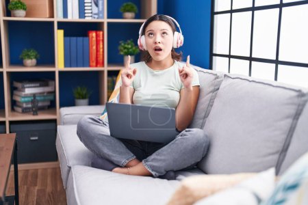 Foto de Hispanic young woman using laptop at home wearing headphones amazed and surprised looking up and pointing with fingers and raised arms. - Imagen libre de derechos