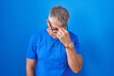Foto de Hispanic man with grey hair standing over blue background tired rubbing nose and eyes feeling fatigue and headache. stress and frustration concept. - Imagen libre de derechos